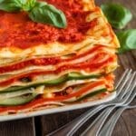 A layered vegan lasagna with tomato sauce, cashew ricotta, and vegetables, garnished with basil on a rustic table.