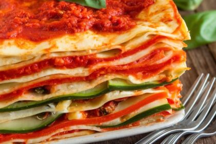 A layered vegan lasagna with tomato sauce, cashew ricotta, and vegetables, garnished with basil on a rustic table.
