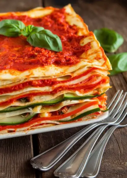 A layered vegan lasagna with tomato sauce, cashew ricotta, and vegetables, garnished with basil on a rustic table.