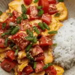 A plate of Chinese tomato and egg stir-fry with scrambled eggs and ripe tomatoes, garnished with green onions, served with rice.
