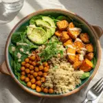 A colorful, vibrant Buddha bowl in a rustic ceramic dish, featuring fluffy quinoa, golden crispy chickpeas, roasted sweet potatoes, bright green avocado slices, and fresh baby spinach. The dish is topped with sesame seeds, microgreens, and a creamy lemon-tahini drizzle. The scene is set on a natural wooden table with a linen napkin, warm sunlight filtering through the window, and a glass of fresh lemon water in the background.