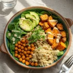 A colorful, vibrant Buddha bowl in a rustic ceramic dish, featuring fluffy quinoa, golden crispy chickpeas, roasted sweet potatoes, bright green avocado slices, and fresh baby spinach. The dish is topped with sesame seeds, microgreens, and a creamy lemon-tahini drizzle. The scene is set on a natural wooden table with a linen napkin, warm sunlight filtering through the window, and a glass of fresh lemon water in the background.