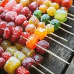Colorful Tanghulu skewers with candied strawberries and grapes on a wooden table.