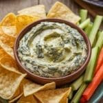 Creamy Spinach and Artichoke Dip Recipe 40 A bowl of creamy spinach and artichoke dip with tortilla chips and vegetable sticks on a wooden table.