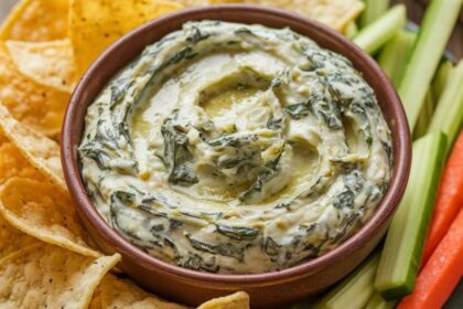 Creamy Spinach and Artichoke Dip Recipe 13 A bowl of creamy spinach and artichoke dip with tortilla chips and vegetable sticks on a wooden table.