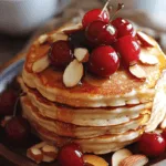 A stack of golden, fluffy cherry almond pancakes on a rustic wooden plate, topped with fresh cherries, sliced almonds, and a drizzle of honey. Soft natural lighting, a cozy breakfast setting with a cup of coffee and a linen napkin beside the plate. The pancakes have a slightly crispy edge and are beautifully layered with melted butter on top.