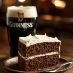 A rich, moist slice of Irish chocolate Guinness cake on a rustic wooden plate, topped with a thick layer of creamy white frosting. A pint of Guinness with a foamy head sits beside it. The background is softly blurred with warm pub lighting, creating a cozy Irish tavern atmosphere.