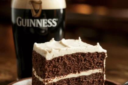 Irish Chocolate Guinness Cake 19 A rich, moist slice of Irish chocolate Guinness cake on a rustic wooden plate, topped with a thick layer of creamy white frosting. A pint of Guinness with a foamy head sits beside it. The background is softly blurred with warm pub lighting, creating a cozy Irish tavern atmosphere.