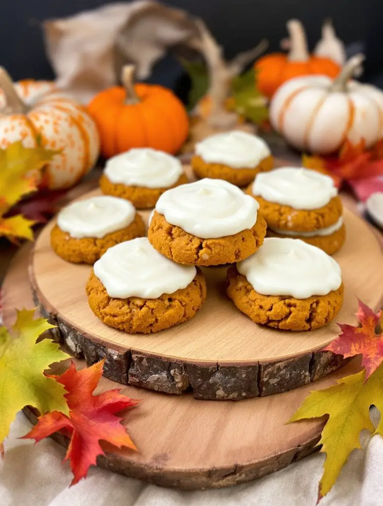 Pumpkin spice cookies with cream cheese frosting on a wooden platter surrounded by autumn leaves and pumpkins.